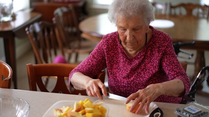 Elderly senior woman slices fruit to make a salad while talking on a phone video call in her kitchen during the afternoon