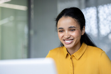 Young multiracial businesswoman smiling at her laptop in a bright modern office, confidently working remotely productive, engaged, and enjoying online learning and virtual meetings