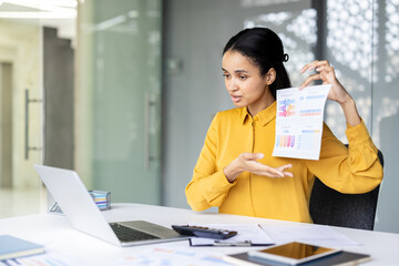 Businesswoman discussing charts and graphs on a printed report while engaging in a video conference from her modern office, analyzing data and sharing information remotely
