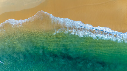 Crystal clear turquoise waves gently wash over smooth golden sand in a serene overhead drone shot of a pristine tropical coastline.