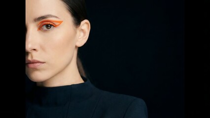 Woman with striking orange eyeshadow and a serious expression, half-lit against a dark background, showcasing bold makeup and a contemplative mood.