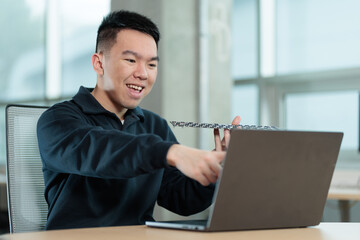 A smiling Asian student holds up an electronic circuit board while pointing at his laptop screen, perfect for STEM education, engineering tutorials, or online technology learning concepts.