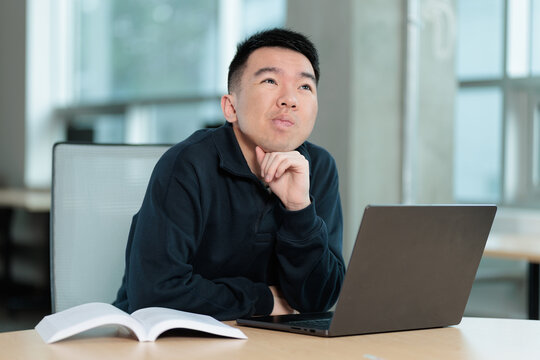 An Asian student looks upward thoughtfully with hand under chin while sitting at a desk with an open textbook and laptop, ideal for study break, brainstorming, or creative thinking concepts.
