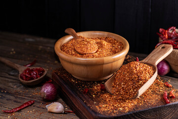 Chili powder in a wooden bowl and with a wooden spoon, along with garlic, shallots, and a mix of Asian spices, are arranged on a wooden table, conveying the concept of spicy food.