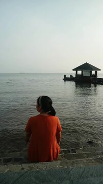 Woman in orange sitting on steps by the water looking out at a pier with a gazebo at dusk.