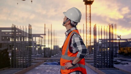 Side View Of Asian Male Engineer Wearing Safety Helmet Looking Around While Standing With Arms Akimbo at Construction Site with Tower Cranes and Building Foundations