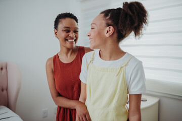 Happy Black mother and teenage daughter laughing together while bonding at home in a bright bedroom.