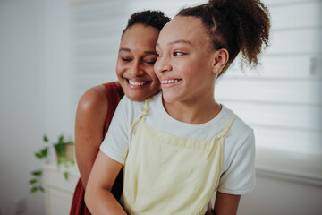 Fototapeta premium Happy African American mother hugging her teenage daughter from behind at home, sharing a joyful moment of love and connection.