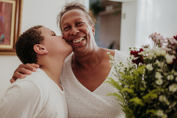 Happy African American grandmother receiving a kiss on the cheek from her grandson while holding a bouquet of flowers at home.