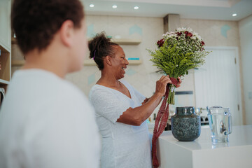 Happy senior African American woman smiling while arranging a beautiful flower bouquet in a modern kitchen with her grandson in the foreground.