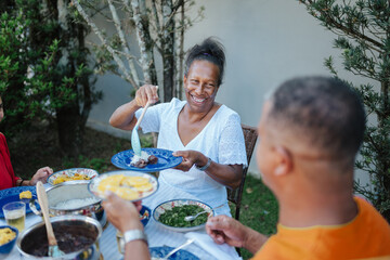 Happy African-Brazilian woman serving traditional feijoada during a family lunch outdoors in a backyard garden.