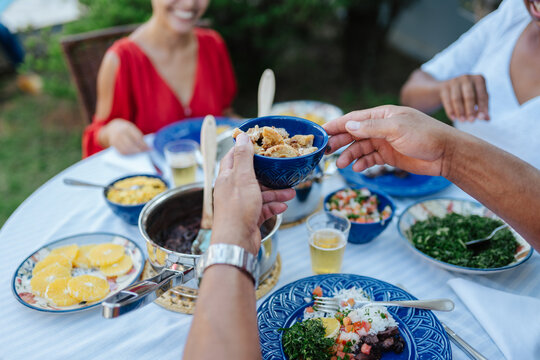 Close-up of hands passing a bowl of pork rinds during a traditional Brazilian feijoada lunch with friends outdoors.