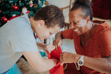 Happy grandmother and grandson opening a red Christmas stocking together at home near a decorated tree during the holiday season.