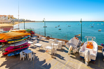 Fishing boats and kayaks near a small outdoor cafe along the sandy La Caleta beach on the Mediterranean Sea coastline of Cadiz, Spain.