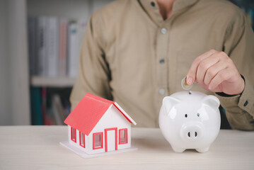 Man putting coin into white piggy bank near house model. Concept of saving money for buying new home, real estate investment, and mortgage financial planning.