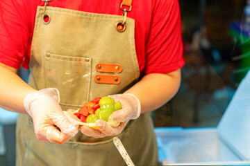 A street food vendor's hands wearing hygienic gloves as they wrap fresh strawberries and green grapes into a soft mochi dough