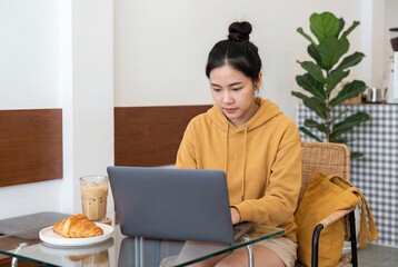 Asian woman in yellow hoodie working on laptop at glass table in cafe with iced coffee