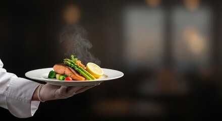 A chef's hand holding a plate of salmon with asparagus and a lemon wedge.