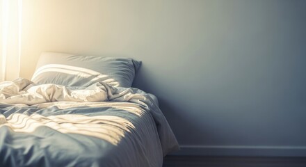 A messy bed with a rumpled blanket and pillows in a bedroom with a light blue wall and a window with sunlight streaming in.