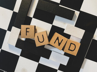 Wooden letter blocks spelling "FUND" are arranged on a black and white checkered board, illustrating capital management, investment pool strategy, and calculated financial resource allocation.
