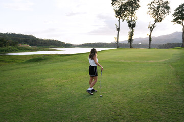 Latin female golfer stands on the green, holding her club, contemplating the next shot amidst a scenic lake and mountains