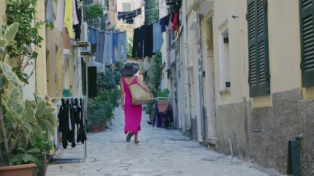 Woman witha hat walks on the street of the ancient Greek city awearing red dress, Corfu Island in Greece, slow motion