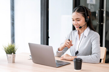 Smiling Asian female customer support agent wearing headset working on laptop in office, providing online service and technical help to clients.