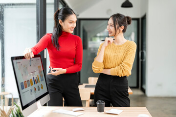 Two professional Asian businesswomen analyzing financial charts and marketing data on computer screen in office.
