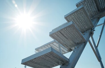 Metal staircase with perforated steps against bright sun and blue sky. Industrial construction and modern architecture create a sense of upward progress.