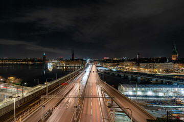 Night long exposure of Stockholm, Sweden, looking across a brightly lit bridge with long-exposure traffic trails leading into the historic city skyline along the waterfront.