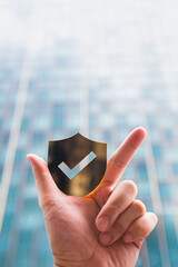 Hand holding shield icon with check mark over blurred modern building background, representing...