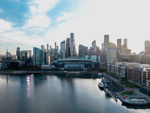 Panoramic aerial view of Melbourne Docklands at sunrise or sunset, highlighting the vibrant city, stadium, and tranquil water.
