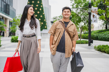 A candid outdoor photograph capturing two Asian friends strolling past retail storefronts. Holding numerous shopping carriers, they engage in animated conversation, reflecting a shared positive experi