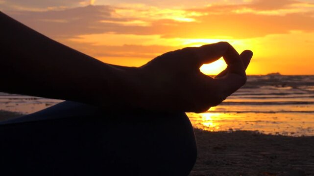 Silhouetted woman's hand in a yoga mudra pose during a beautiful sunset on the beach, creating a serene and mindful atmosphere with golden sun reflections on the calm sea waves