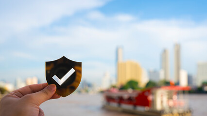 Hand holding a shield with check mark icon over blurred modern cityscape and river view, representing urban security, verified service, trust, data protection, and digital privacy concept.