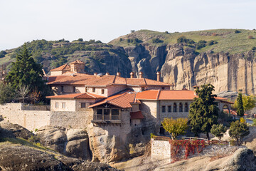 Obraz premium Traditional stone monastery complex with terracotta rooftops sits atop rugged sandstone rocks in Meteora, Greece, surrounded by greenery and dramatic cliffs in warm daylight.