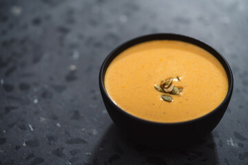 Pumpkin cream soup with pumpkin seeds in a black bowl on concrete background
