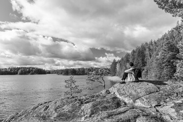 Black and white landscape photograph of a person sitting on a rocky shoreline by a calm lake, surrounded by forest and dramatic clouds, evoking solitude, contemplation and connection with nature