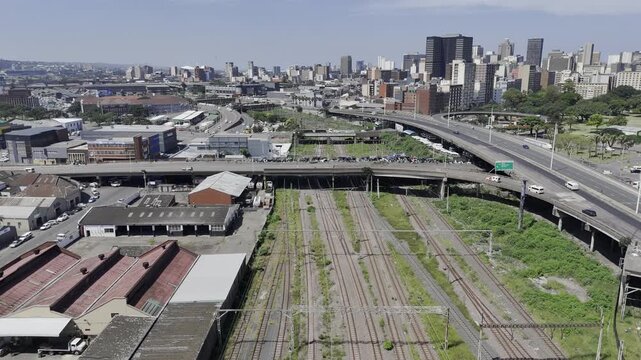 Drone lowers over train tracks that cross under bridge with informal settlement on sunny day in Berea, Durban, South Africa