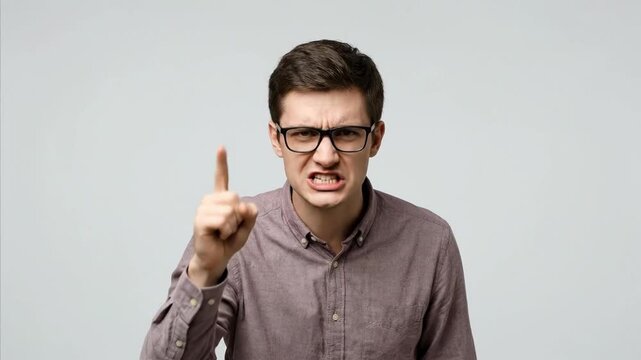 A young adult male with glasses and a casual collared shirt is captured in a close-up shot, intensely pointing his finger directly at the viewer. His mouth is open in a shouting or yelling gesture, co