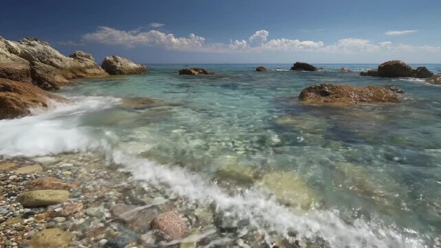 Immerse yourself in the serene beauty of a pristine coastal landscape on a vibrant sunny day. Crystal clear turquoise-blue waters flow gently over a pebble-strewn seafloor, revealing the intricate det