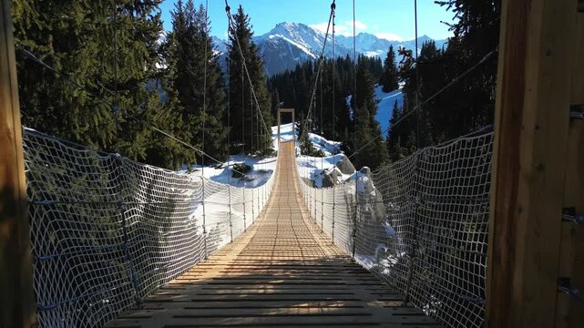 First-person perspective crossing a long wooden suspension bridge over a snowy valley. Scenic mountain landscape with pine trees
