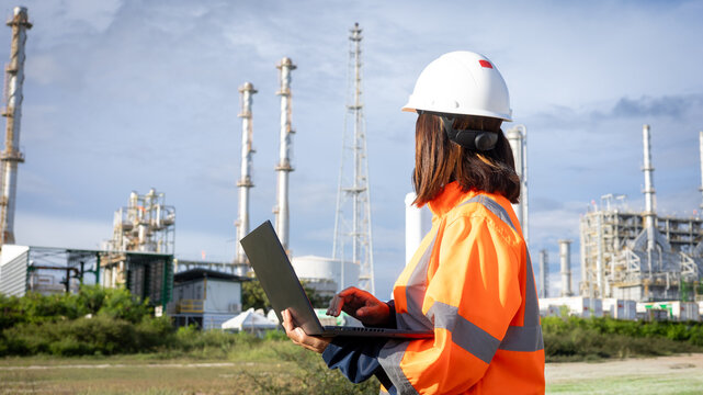 A worker stands outside at an industrial site. The person uses a laptop while wearing a hard hat and safety gear. Towers and equipment are seen in the background