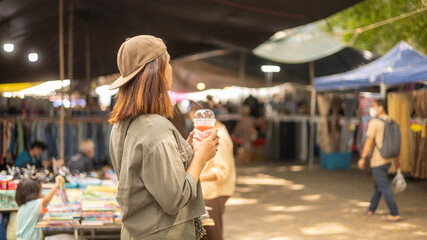 A woman stands with a drink in her hand looking at vendors in a busy market. Many people walk around shopping. Tents and stalls surround the area. It is a sunny day © Happy Photo