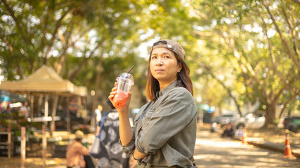 Fototapeta premium A young woman stands in a park holding a drink. She gazes thoughtfully while wearing a cap and casual clothing. Trees and people are visible in the background