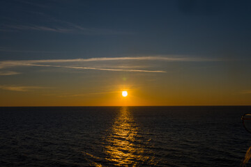 Fototapeta premium The sun sets low above the calm Adriatic Sea, casting a golden reflection across the rippling water. Subtle clouds and a deepening blue sky create a peaceful, minimal seascape.