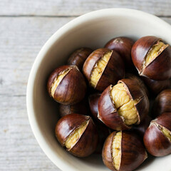 Top view of roasted chestnuts with cracked shells served in a white ceramic bowl on a light gray wooden surface