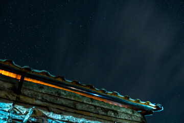 Night sky over the old cabin lit with a warm glow as stars twinkle above a wooden roof