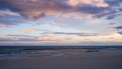 Fototapeta premium Soft pastel clouds stretch across the evening sky above a deserted sandy beach in Agde, with gentle waves and a row of yellow pedal boats adding subtle detail to the tranquil coastal scene.