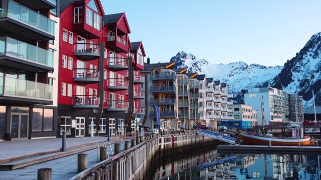Footsteps echo along the harbor as morning light shines on red and gray buildings. The water is still, showing clear reflections of the structures. Boats are anchored nearby.
